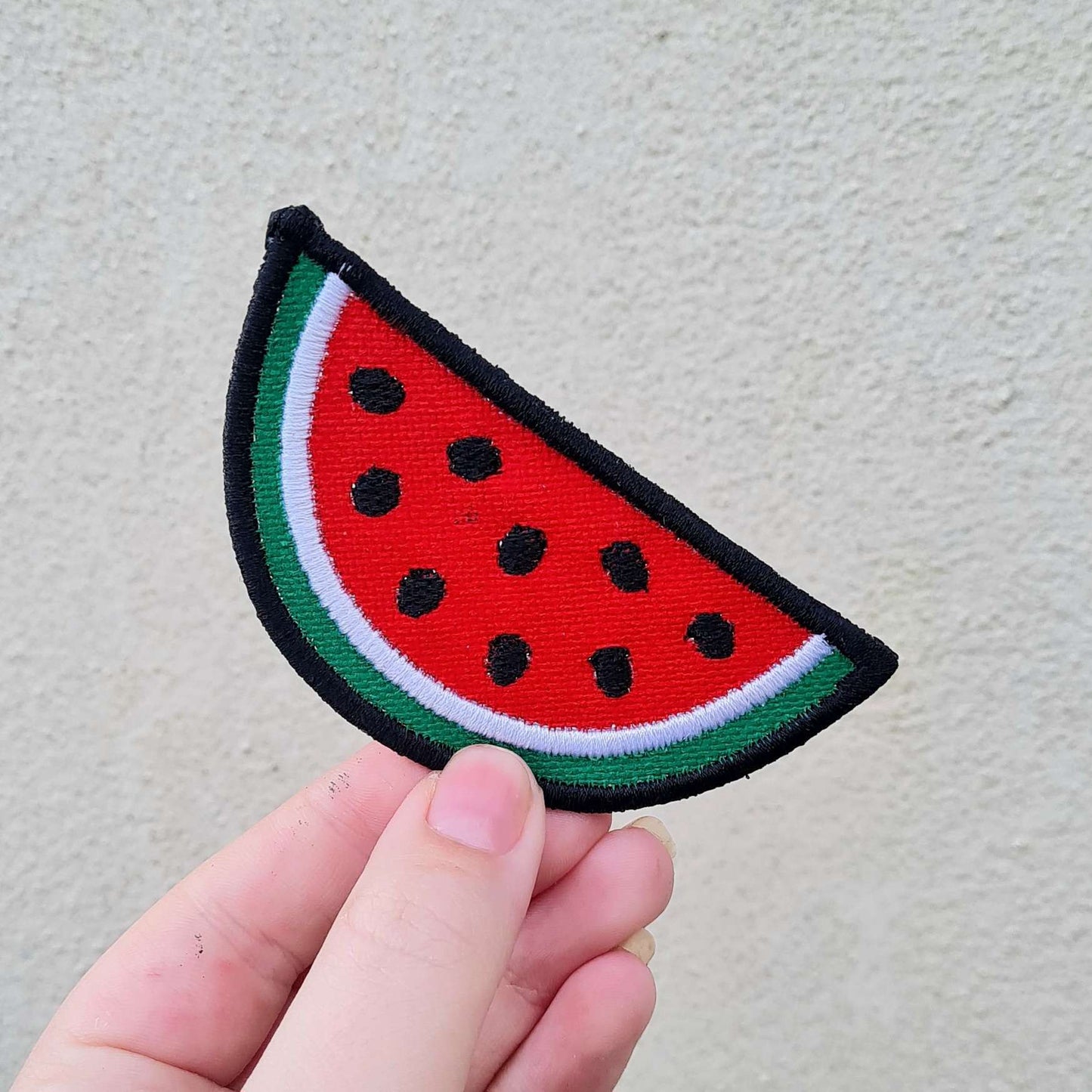 A watermelon patch held against a cream background. The patch is a semi circle edged in black, followed by green, then white, with the rest of the patch being filled in red with black seeds embroidered on top.