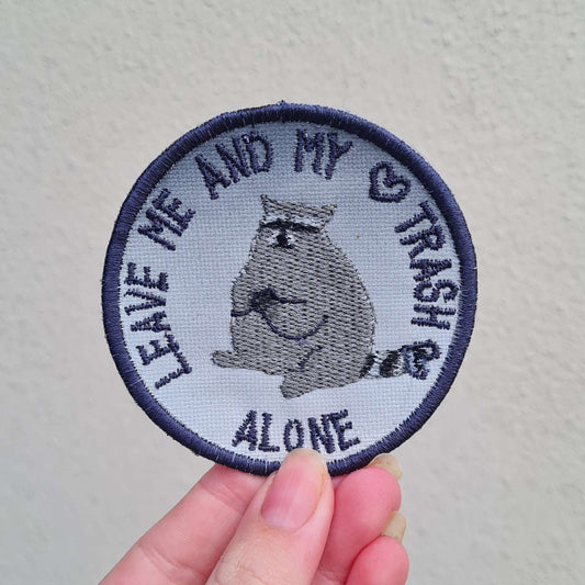 A circular patch held up against a cream background. The patch is edged in dark grey with text in the same colour saying 'LEAVE ME AND MY ❤️ TRASH ❤️ ALONE'. The background of the patch is a medium grey at the top, fading to a lighter grey at the bottom. In the centre of the patch is a round-looking raccoon sitting down, back legs splayed apart, with its front legs clutched together, holding something.